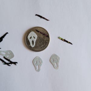 May include: A close-up of a US quarter with a ghost face mask sticker on top. There are two more ghost face mask stickers, a black knife, and a floral patterned knife on a white surface.