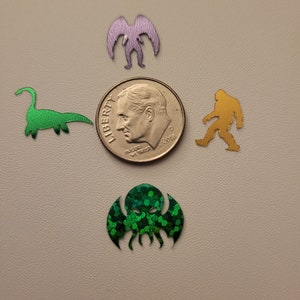 May include: Five different monster silhouettes cut from metallic paper. The silhouettes are of a green dinosaur, a purple alien, a gold Bigfoot, a green Cthulhu, and a silver coin with the text "LIBERTY" and "IN GOD WE TRUST".