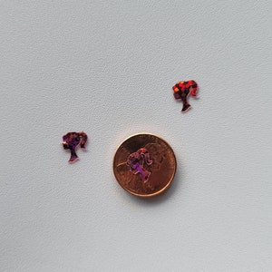 May include: Two small, glittery, red and purple, cutout images of a woman's head with a ponytail. The images are next to a US penny.