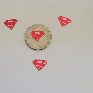 May include: Three red Superman logos cut from paper, with one logo centered above a US quarter.