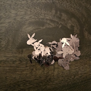 May include: A pile of rose gold bunny shaped metal charms. The charms are scattered on a dark brown wooden surface.