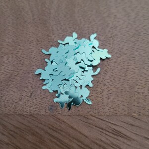 Pu&ograve; includere: Un mucchio di coriandoli metallici blu-verde a forma di foglie. I coriandoli sono sparsi su una superficie di legno marrone.