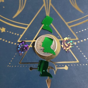 May include: A silver US coin with the text "LIBERTY" and "IN GOD WE TRUST" on it. The coin is surrounded by four green glitter shapes, including a silhouette of a person wearing a hat.