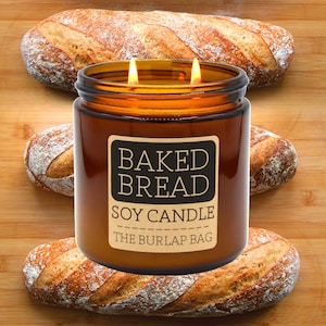 May include: A brown glass candle with a label that reads "Baked Bread Soy Candle The Burlap Bag". The candle is surrounded by three loaves of bread.
