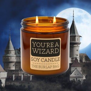 May include: A brown glass soy candle with a label that reads "You're a Wizard Soy Candle The Burlap Bag". The candle is lit and has two flames.