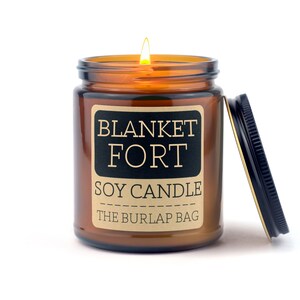 Puede incluir: Una vela de vidrio marr&oacute;n en un frasco con una etiqueta de papel marr&oacute;n que dice "BLANKET FORT SOY CANDLE THE BURLAP BAG". La vela est&aacute; encendida y la tapa est&aacute; abierta.