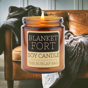 Puede incluir: Una vela de vidrio marr&oacute;n con una etiqueta que dice "Blanket Fort Soy Candle The Burlap Bag". La vela est&aacute; encendida y se encuentra sobre un sof&aacute; de cuero marr&oacute;n con una manta sobre &eacute;l.