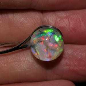 May include: A silver earring with a round, iridescent opal stone. The opal displays a vibrant play of color, including green, red, and blue hues. The earring's design is simple, focusing on the beauty of the gemstone.