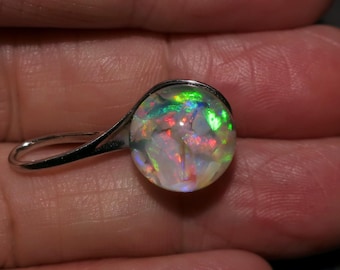 May include: A silver earring with a round, iridescent opal stone. The opal displays a vibrant play of color, including green, red, and blue hues. The earring's design is simple, focusing on the beauty of the gemstone.