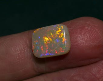 May include: A white opal gemstone with a rectangular shape and a rainbow of colors, including red, orange, yellow, green, blue, and purple.