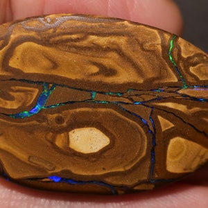 Könnte beinhalten: Ein brauner und schwarzer Opal-Cabochon mit blauen und grünen irisierenden Blitzen. Der Opal hat eine natürliche, organische Form.
