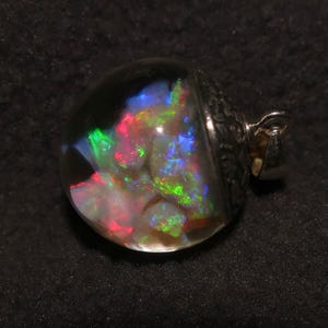 May include: A clear, round pendant with a silver-coloured metal setting. The pendant contains a colourful, iridescent stone with flashes of red, green, and blue. The stone is encased in a clear, rounded glass or resin. The pendant has a small loop for a chain.