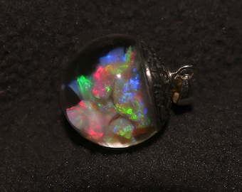 May include: A clear, round pendant with a silver-colored metal setting. The pendant contains a colorful, iridescent stone with flashes of red, green, and blue. The stone is encased in a clear, rounded glass or resin. The pendant has a small loop for a chain.