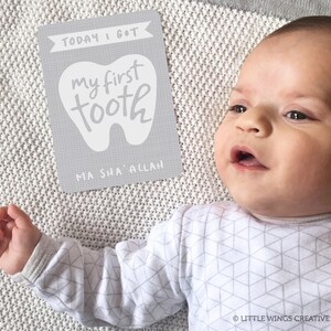 May include: A gray card with the text "TODAY I GOT my first tooth MA SHA' ALLAH" in white lettering. A baby is lying on a white blanket with a gray geometric pattern.