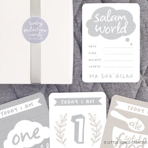 May include: A set of gray baby milestone cards with a white background. The cards feature a variety of designs, including a cloud with the text "Salam world", a number one with leaves, and a spoon and bowl with the text "I ate solid food". The cards also include the text "Today I am" and "Ma Sha' Allah".
