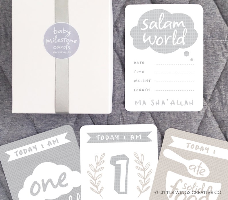 May include: A set of gray baby milestone cards with a white background. The cards feature a variety of designs, including a cloud with the text "Salam world", a number one with leaves, and a spoon and bowl with the text "I ate solid food". The cards also include the text "Today I am" and "Ma Sha' Allah".