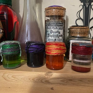May include: Four small glass bottles with cork tops and colorful liquids inside. Each bottle is wrapped with twine in different colors: green, purple, orange, and red. The bottles are arranged on a wooden shelf.