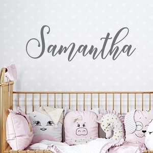 May include: A gold crib with a pink and white bedding set. The crib is decorated with pillows featuring cartoon animal faces. The wall behind the crib has a gray cursive name "Samantha" written on it.