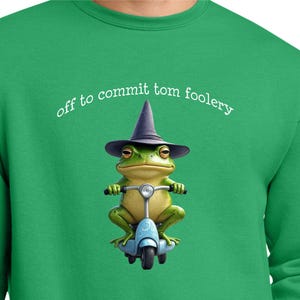 May include: Green sweatshirt with the phrase "off to commit tom foolery" in white. The graphic features a frog wearing a witch hat and riding a blue scooter. The frog is green and yellow.