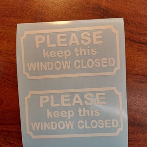 May include: Two white vinyl decals on a light blue backing sheet. Each rectangular decal features a decorative border and the text "PLEASE keep this WINDOW CLOSED" in a clean, sans-serif font, designed for application to a glass surface.