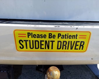 May include: A rectangular yellow bumper sticker with a thin red border. The text reads "Please Be Patient" in black, followed by "STUDENT DRIVER" in large, bold black capital letters. Two red horizontal lines flank the top text.