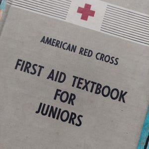 May include: A light gray hardcover book titled American Red Cross First Aid Textbook for Juniors. The cover features a red cross symbol at the top, black text, and horizontal line details. The book has a textured fabric-like material finish.