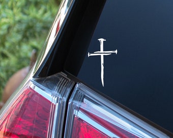 May include: A white vinyl decal featuring a cross design composed of two intersecting iron nails. The decal is applied to the dark glass surface of a vehicle window, displaying a sharp, high-contrast graphic of the nail-shaped cross symbol.
