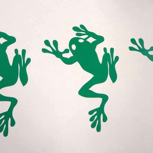 May include: A set of three identical green frog decals arranged in a horizontal row. Each frog is depicted in a climbing or leaping pose with splayed toes and a rounded head, featuring a solid green silhouette against a plain white background.