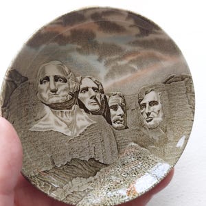 May include: A small, round ceramic dish featuring a depiction of Mount Rushmore. The image shows the faces of four U.S. presidents carved into a mountain, with a cloudy sky background. The dish has a brown and beige color scheme.