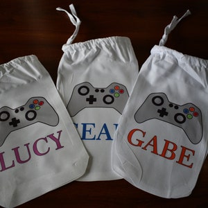 May include: Three white drawstring bags with a gray video game controller design. The bags are personalized with the names "Lucy", "Leah", and "Gabe".