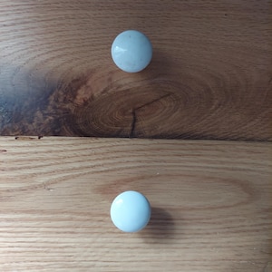 May include: Two smooth, round, white ceramic knobs are attached to a light brown wooden surface with a visible grain pattern. The knobs are positioned vertically, one above the other, against the natural wood texture of the furniture panel.