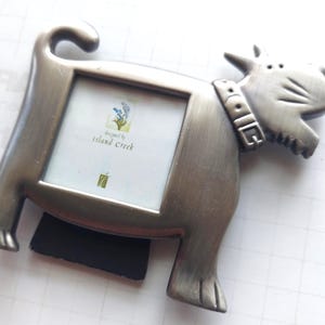May include: A silver-colored dog-shaped picture frame with a rectangular opening. The frame has a magnet on the back and the text "designed by island creek" is visible on the insert. The dog has a collar that says "DOG".