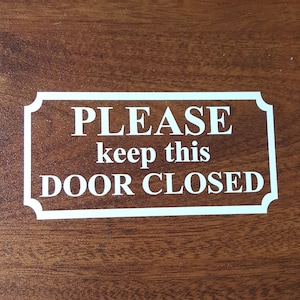 May include: A white rectangular vinyl decal with rounded corners featuring the text PLEASE keep this DOOR CLOSED in a bold, serif font. The decal is applied to a smooth, medium-brown wood grain surface with a visible horizontal pattern.