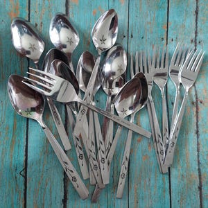 May include: A collection of stainless steel flatware including several spoons and forks. Each piece features a polished finish and a slender handle decorated with a delicate etched floral vine pattern. The metal surfaces reflect light.