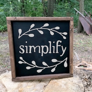 May include: A black framed sign with the word "simplify" written in white lettering. The sign is decorated with a white floral design.