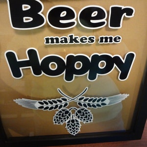 May include: A framed sign with the text "Beer makes me Hoppy" in black letters with white outlines. The sign has a brown background and a black frame. There are two white wheat stalks with black outlines and two black hops with white outlines at the bottom of the sign.