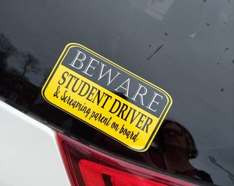 May include: A rectangular yellow bumper sticker with a black border and black text that reads BEWARE STUDENT DRIVER & screaming parent on board. The sticker is applied to the rear window of a vehicle above a red taillight assembly.
