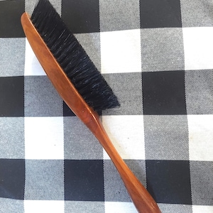 May include: A wooden clothes brush with a long handle and black bristles. The brush has a curved design and a polished finish. The handle is a warm brown color, and the bristles are densely packed.