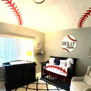 May include: A nursery decorated with a baseball theme. The ceiling has red baseball stitching decals. A crib with a red, white, and blue baseball themed bedding set. A white armchair with a gray cushion. A wooden sign with the text "Reilly Quinn" on a white baseball with red stitching.