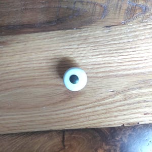 May include: A small, white, round cabinet knob with a centered, dark gray hole. The knob is set against a backdrop of light brown wooden planks.