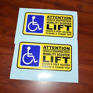 May include: Two rectangular stickers with a blue wheelchair symbol and the text "ATTENTION VEHICLE EQUIPPED WITH MOBILITY SCOOTER LIFT LEAVE 8 FEET ACCESS PLEASE & THANK YOU" in black and yellow.