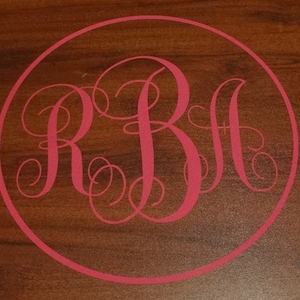 May include: A pink monogram with the letters "RBH" in a circle on a brown wooden surface.