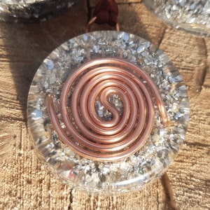 May include: A round, clear pendant with a copper wire spiral design embedded inside. The pendant is filled with silver glitter and has a brown cord attached.