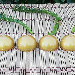 Peut inclure: Quatre objets dorés en forme de dôme sont disposés en rangée sur un tapis en bambou. Les objets ont une surface lisse et brillante. Des frondes de fougères vertes sont placées autour des objets, ajoutant un élément naturel à la composition.