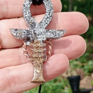 May include: Ankh pendant with a silver-colored top loop and crossbar, and a gold-colored lower section. The pendant is suspended from a black cord. The ankh is a symbol of life and immortality, often associated with ancient Egyptian culture.