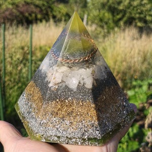 Könnte beinhalten: Ein pyramidenförmiges Objekt aus Kunstharz mit Schichten aus silbernen und goldenen Metallflocken an der Basis, einem Mittelteil aus klaren Quarzkristallen und einem Oberteil mit einem gewickelten Kupferdraht in durchscheinendem grünem Harz.