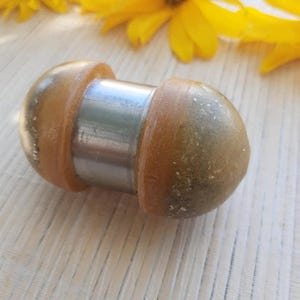 May include: A bottle stopper with a stainless steel centre and two rounded, amber-coloured ends. The stopper is lying on a white surface with yellow flowers in the background.