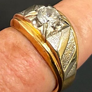 May include: A two-tone ring featuring a central round clear gemstone set in a white metal prong setting. The band is crafted from polished yellow gold and accented with textured, triangular white metal panels on the sides of the central stone.