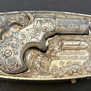 以下が含まれることがあります： マテル社製のレミントン・デリンジャー1867ピストルがエンボス加工された、銀色の長方形の金属製ベルトバックル。表面には複雑なスクロール模様と花柄が施されており、弾丸のアイコンと「REMINGTON DERRINGER 1867 By Mattel」という文字が刻まれています。
