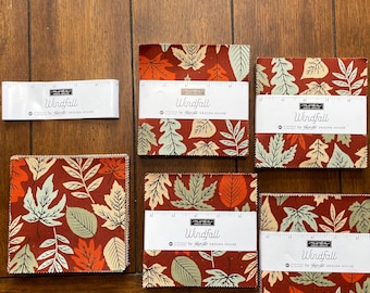 May include: Five stacks of square fabric pieces and one fabric label strip, all featuring a Windfall pattern by Moda Fabrics. The fabric displays a repeating autumn leaf motif in shades of rust orange, cream, and sage green on a dark red background.